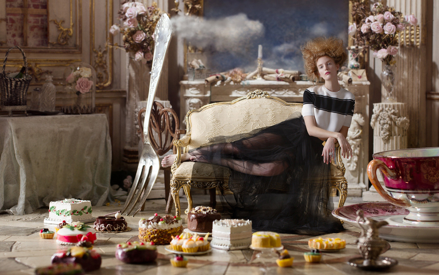A blonde-haired fashion model reclining on an Louis XVI sofa in an elegant baroque interior surrounded by a display of pastries and cakes on the tile floor and also a giant sterling silver fork and giant antique teacup.