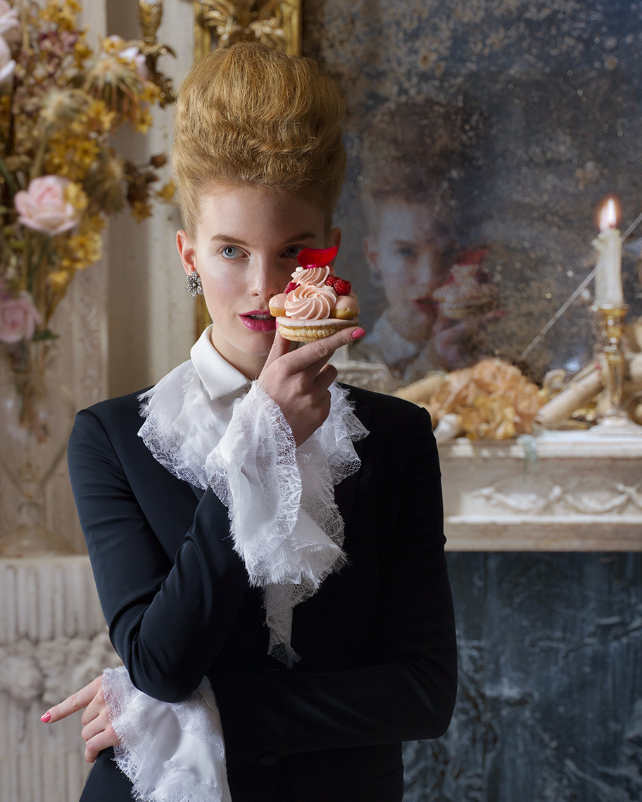 Blonde fashion model with “Marie Antoinette” hair style in Versace dark jacket with white shirt with ruffled collar and sleeves holding a fancy French pastry.