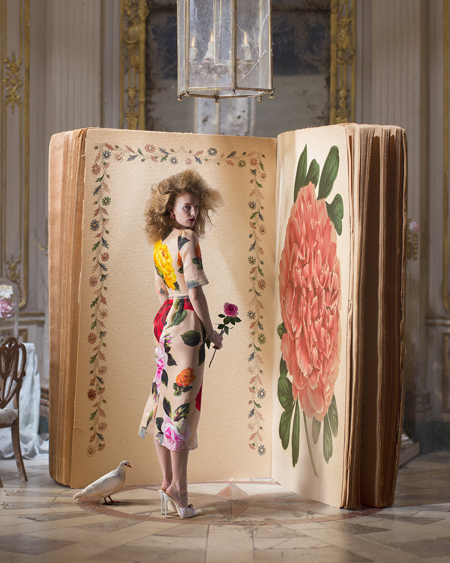 Fashion model with blonde hair like a lion’s mane wearing an elegant Dolce & Gabbana spring floral dress standing in front of a giant book open to a vintage floral illustration in an elegant interior.