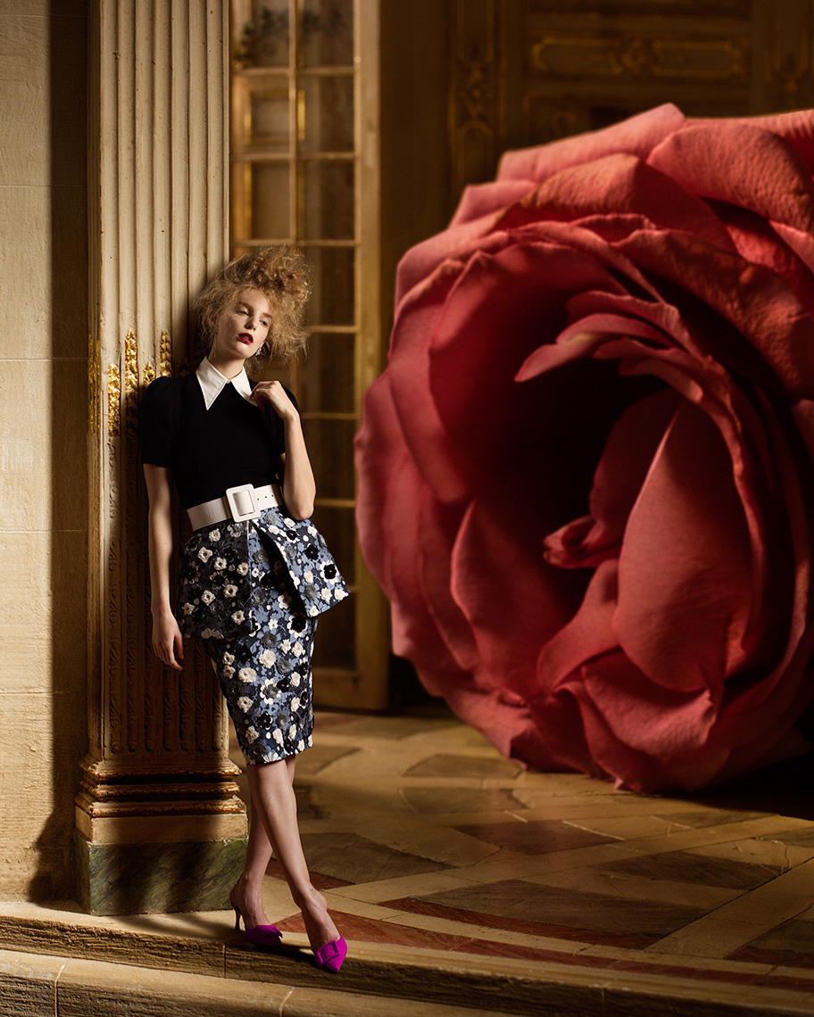 Elegant fashion model wearing stylish clothes leans against a column in a Baroque interior with a gigantic, larger than life red rose in the background.