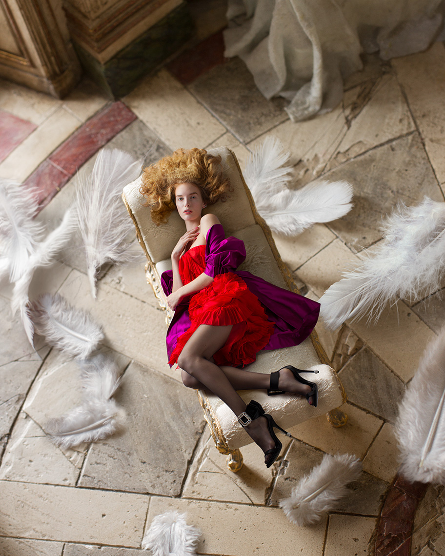 Bird’s eye view of blonde fashion model in black heels and revealing red dress reclining on antique chaise lounge, surrounded by giant feathers on the elegant tile floor.