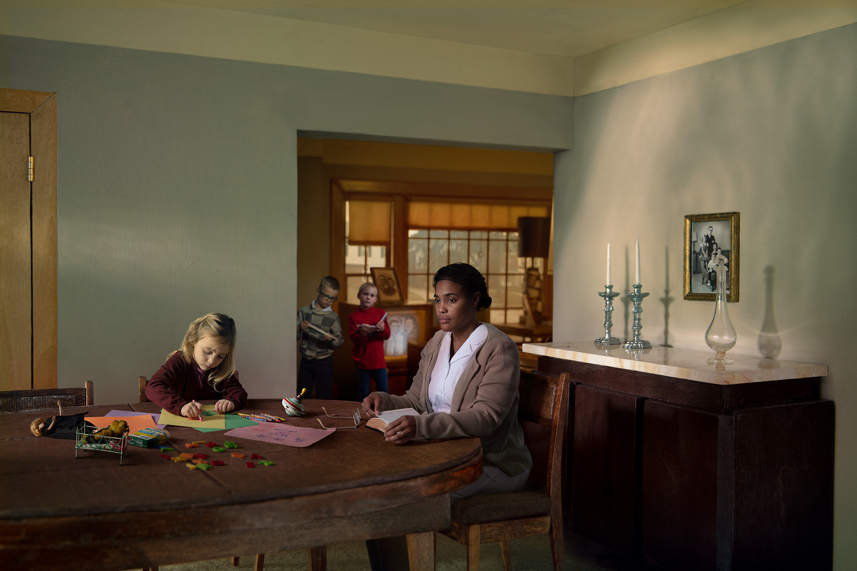 Staged photomontage of African-American nanny lost in thought as young girl draws and boys play in front of TV in 1960s’ suburban interior re-creation.