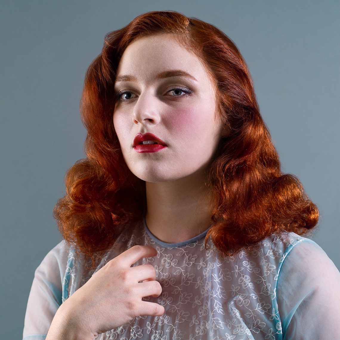 Head & shoulders beauty portrait of a pretty, fair, blue-eyed young woman with shoulder-length wavy red hair, bright red lipstick, and a sheer  light blue silk short-sleeved blouse