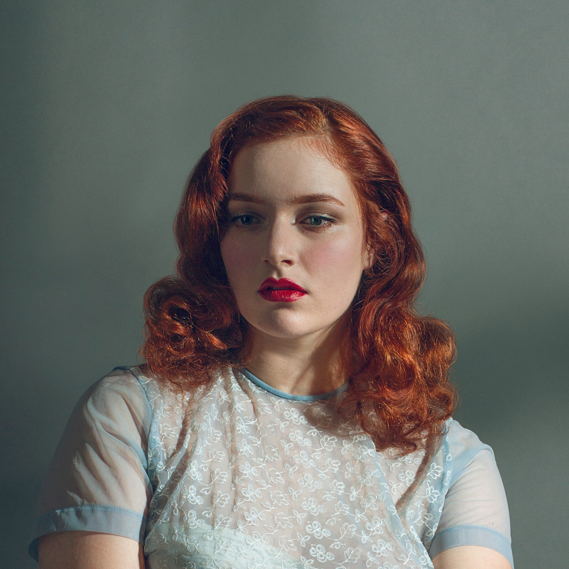 Head & shoulders beauty portrait of a pretty, fair, blue-eyed young woman with shoulder-length wavy red hair, bright red lipstick, and a sheer light blue silk short-sleeved blouse.