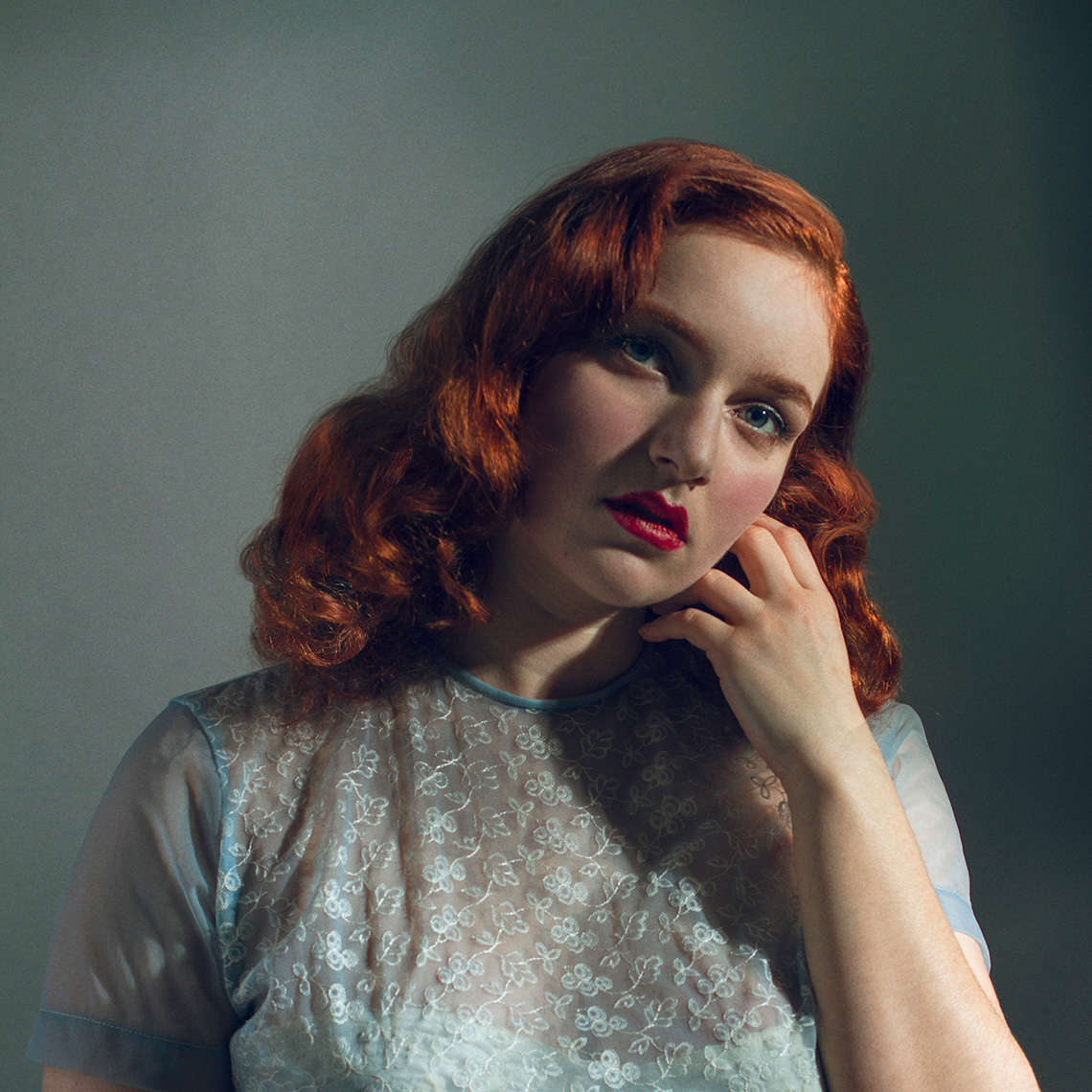 Retro-styled head & shoulders beauty portrait of a pretty, fair, blue-eyed young woman with shoulder-length wavy red hair, bright red lipstick, and a sheer light blue silk short-sleeved blouse.
