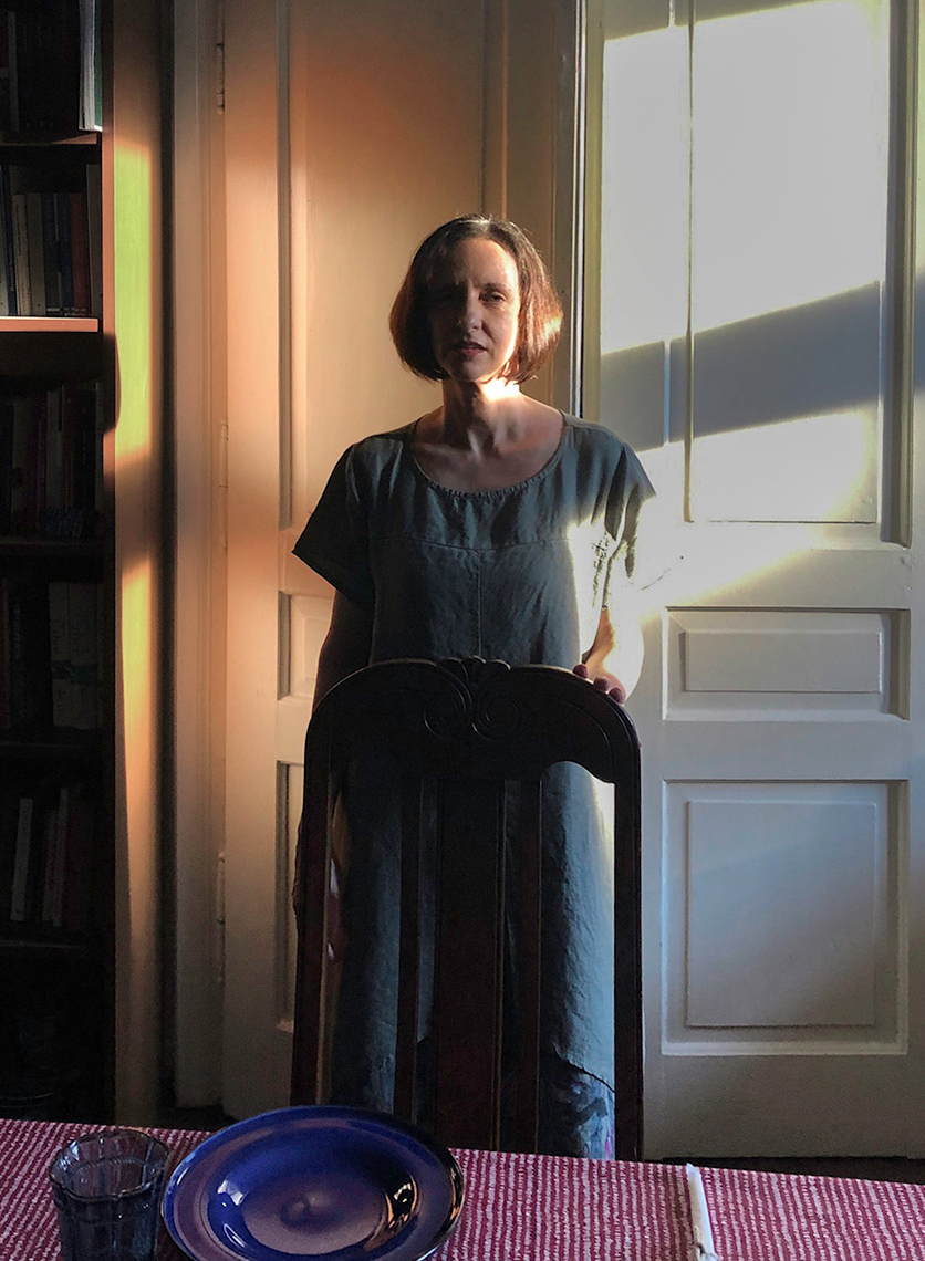 Portrait of a middle-aged woman standing behind a dining room chair and place setting, one side of her flooded with light from the out of frame adjacent window.