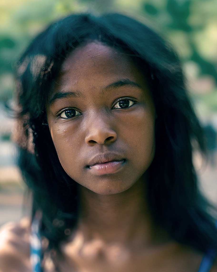 Closeup portrait of a pretty, African-American adolescent girl with shoulder-length hair against a background of soft-focus sun dappled foliage.