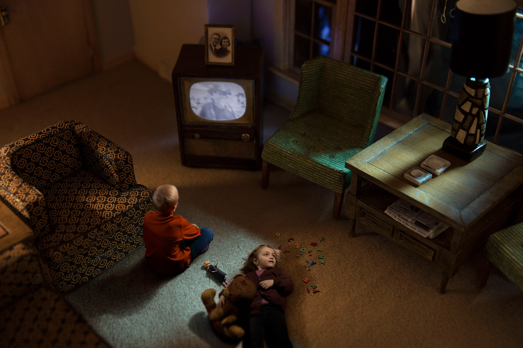 1960s re-creation of brother and sister in suburban living room in the evening, the boy watching a western on TV and the girl daydreaming on the floor with her teddy bear.