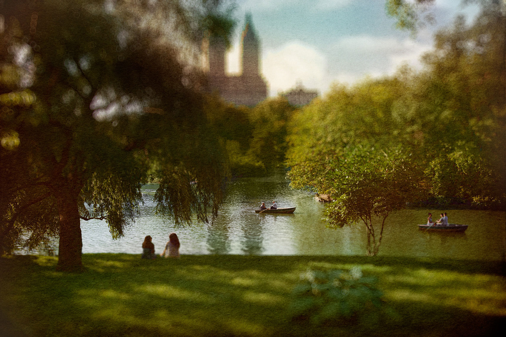 Quiet afternoon in Central Park with two women sitting on sun-dappled grass under a Willow tree with row boats on the lake and an apartment building in the distance.