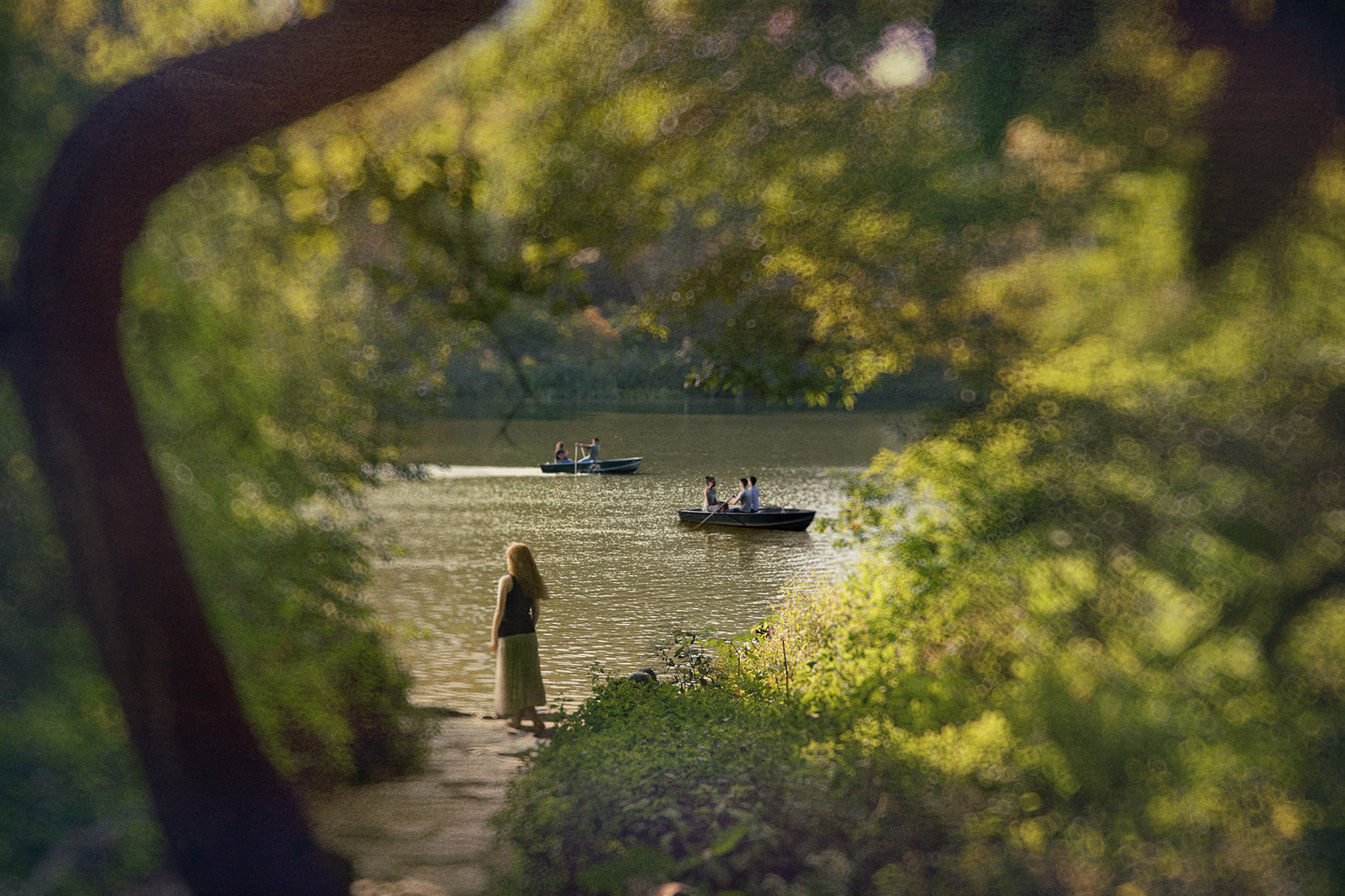 Framed by the lush foliage of soft focus trees, a young woman with long, red hair stands at the edge of a lake, with people enjoying a leisurely day in rowboats in the middle distance.