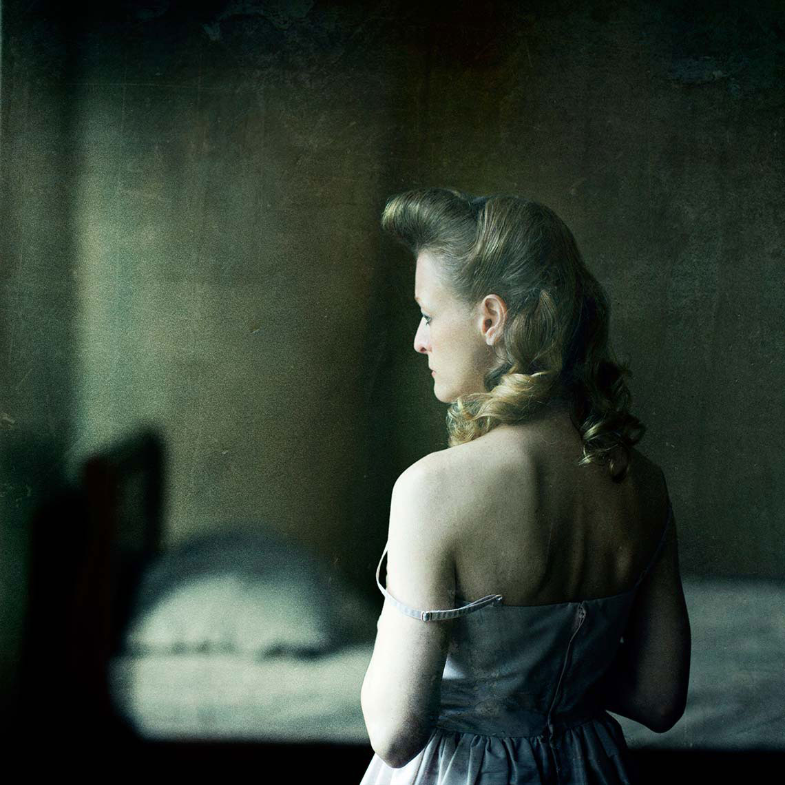 A bare-shouldered melancholic blonde woman with a 1940s’ hairstyle wearing a pale spaghetti-strap dress stands in front of an unmade bed, gazing off-camera in contemplation.