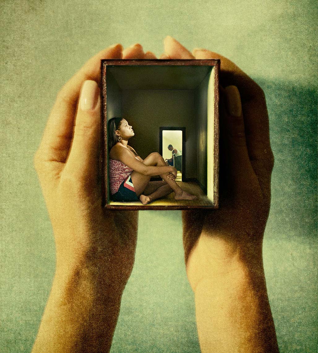 Two hands hold a small box containing  miniature scene of a sad couple, the wife sitting against the wall in the foreground, and the husband sitting on a bed through a doorway in the background.