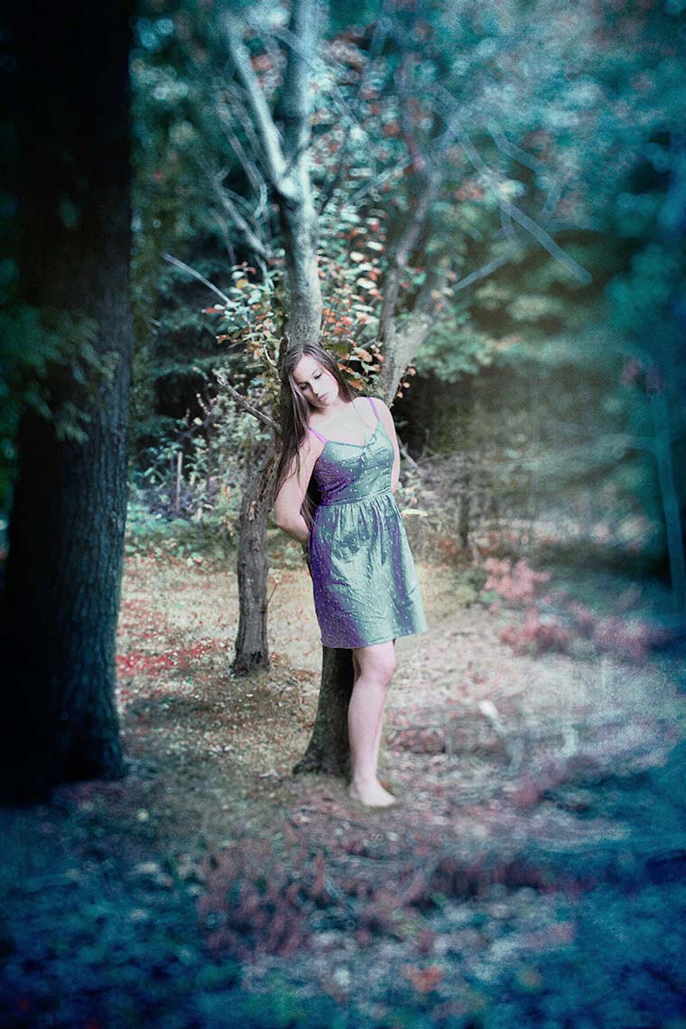 Portrait of a pretty teenage girl in a light blue spaghetti-strap dress, leaning against a tree in the woods, while gazing towards the ground against a soft-focus landscape background.