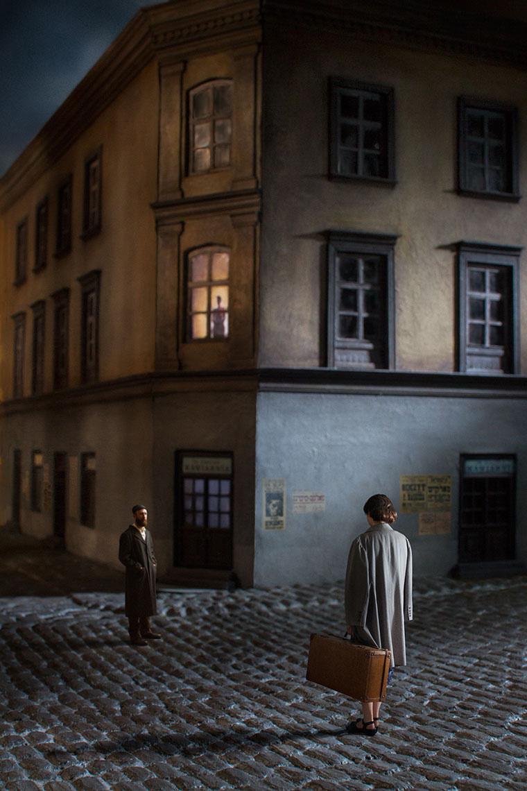 On a shadowy, dark, lonely 1930s Polish cobblestone street at night, a dark-haired young woman wearing an overcoat and carrying a suitcase, her back to the camera, faces a bearded young Orthodox Jewish man across the way as a mysterious figure watches from an apartment window above.