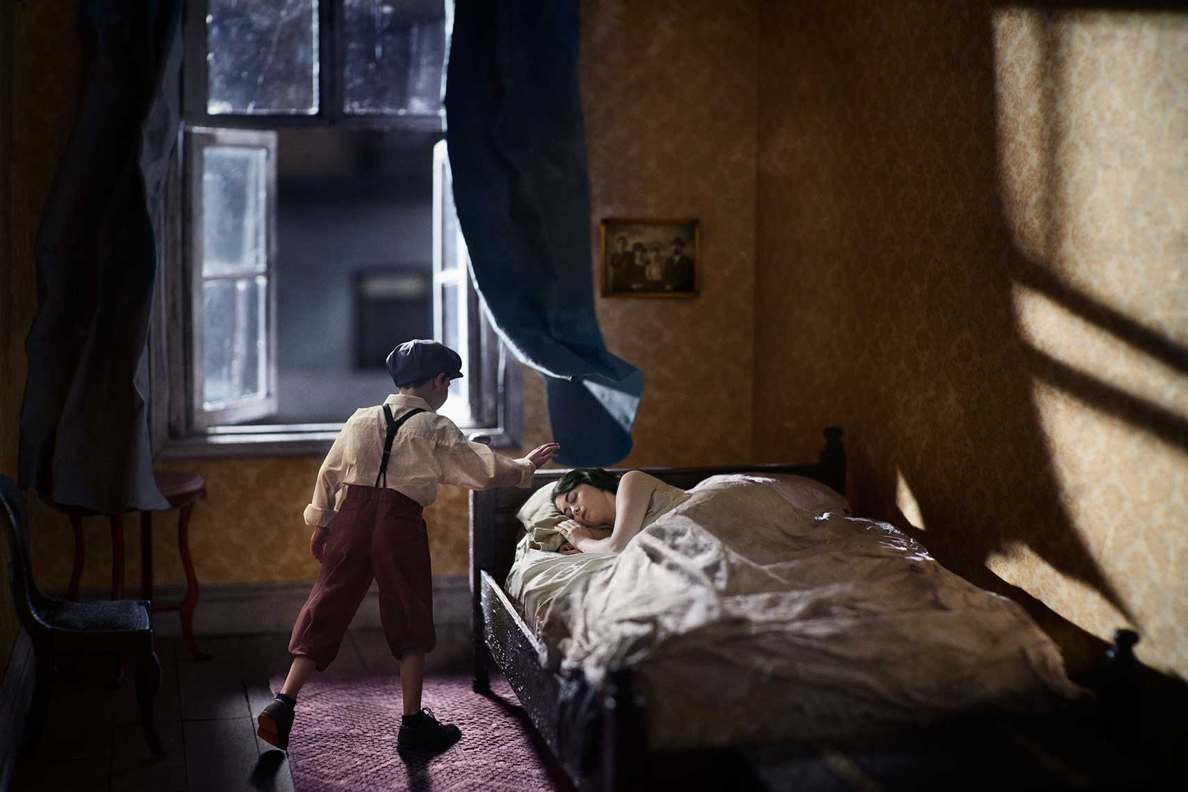 Dramatic painterly photomontage of a 1930s’ European bedroom at night, a young boy in period garb approaches the sleeping woman in bed, the curtain blowing in the breeze from the open window.
