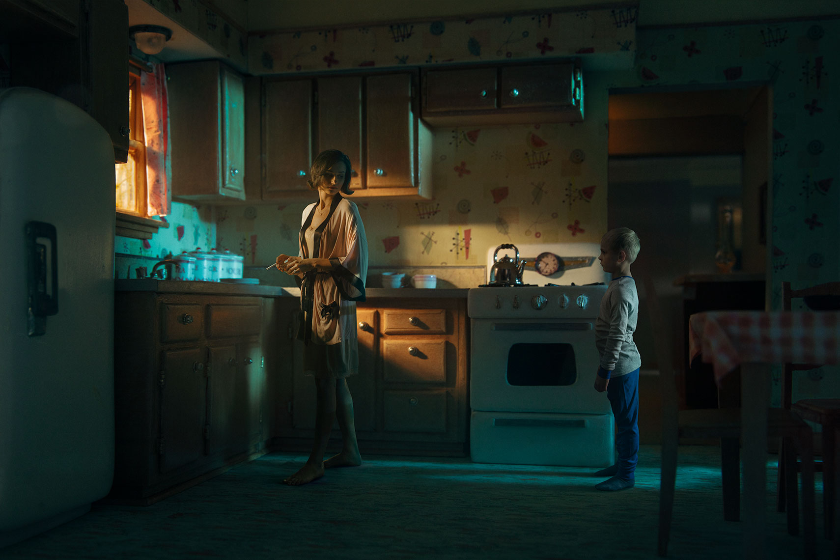 Moody photomontage of mother and young son in night clothes gazing at each other in early morning in sunlit 1960s’ kitchen miniature dollhouse set.