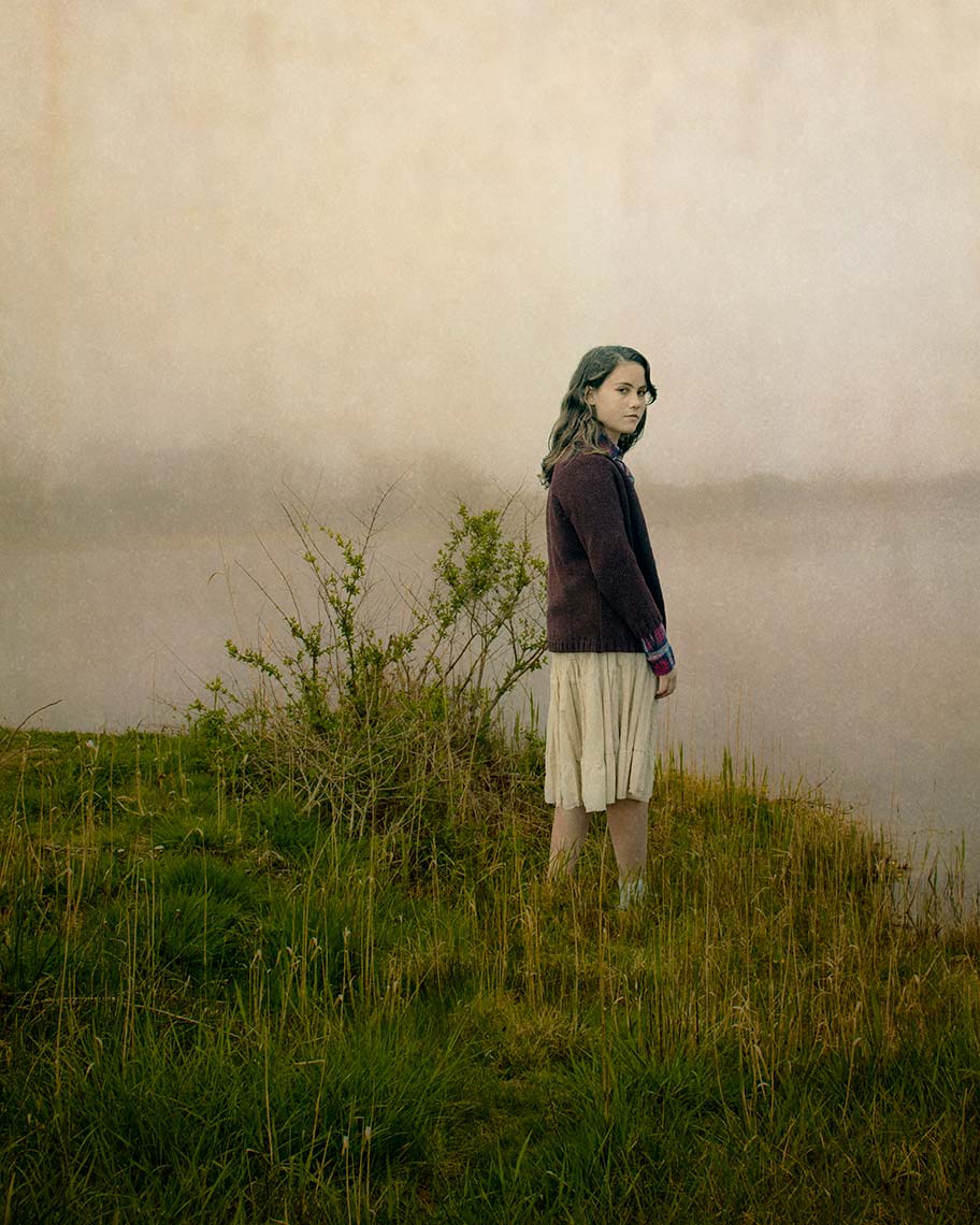 fallon-easthampton-iiA pretty, long-haired teenage girl in a dark sweater and light skirt turns quietly towards the camera, while standing in the long grass at the edge of a pond in a misty, melancholic landscape.