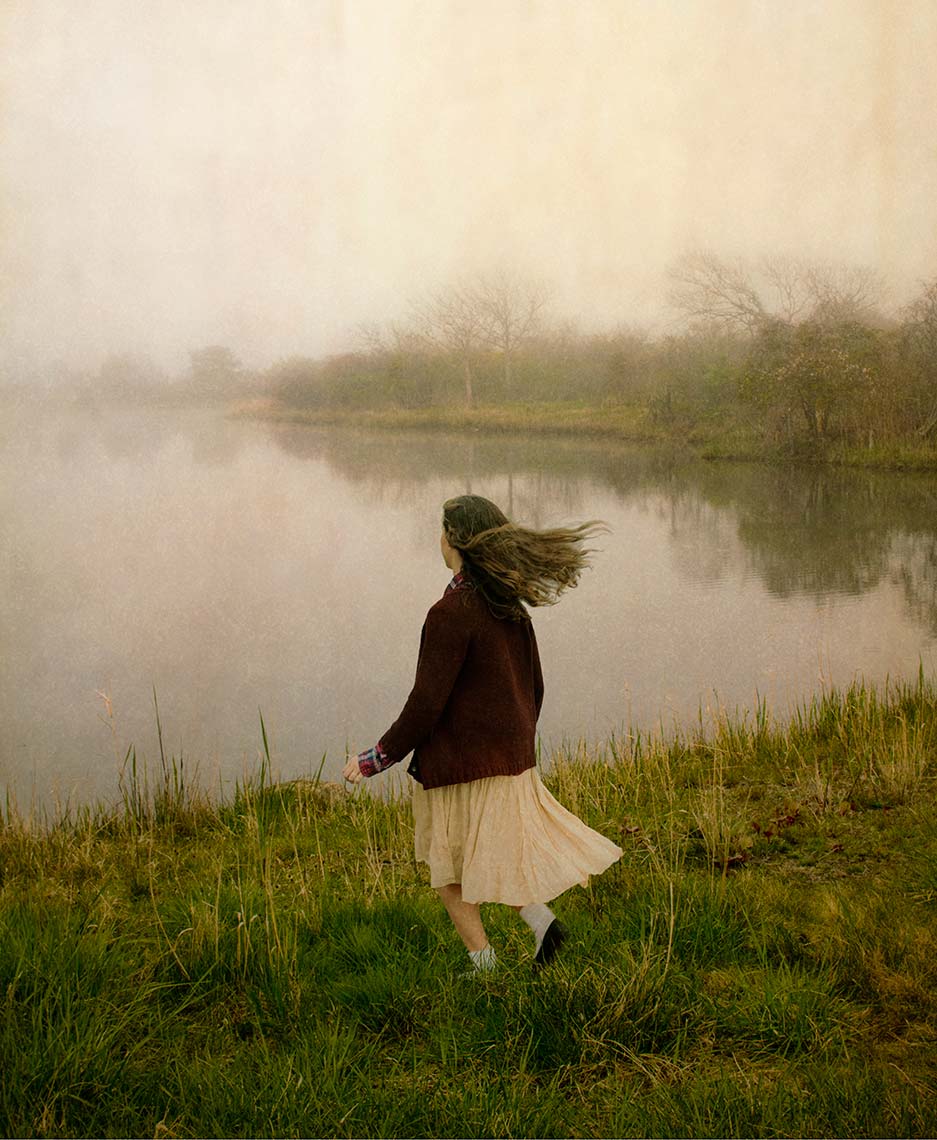 A teenage girl from behind, long brown hair and skirt blowing in the wind, running in the long grass towards a pond in a misty, melancholic landscape.