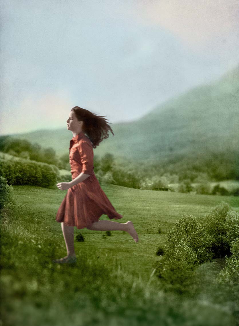 A pretty teenage girl is seen from the side, long brown hair and red dress blowing in the wind, running in the long grass against a backdrop of green, rolling hills.