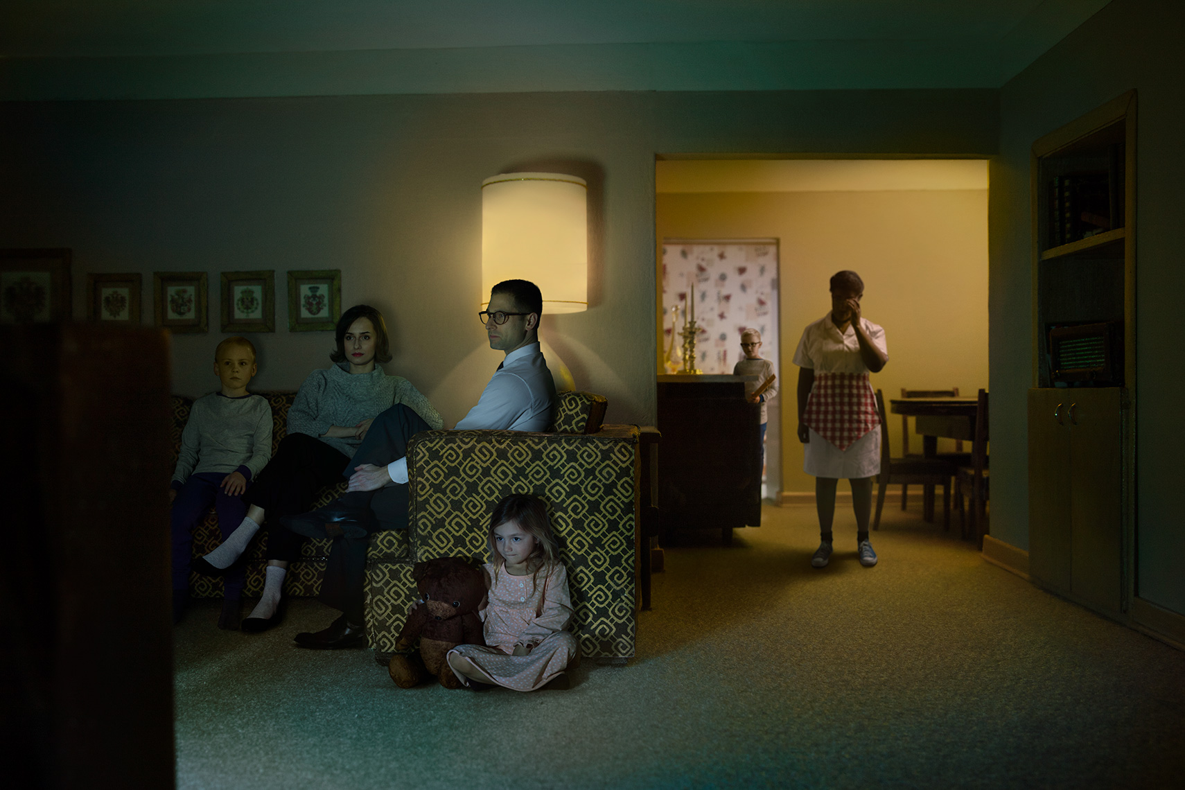 Re-creation of 1960s white suburban family watching TV in the evening while African-American housekeeper looks on in the background.