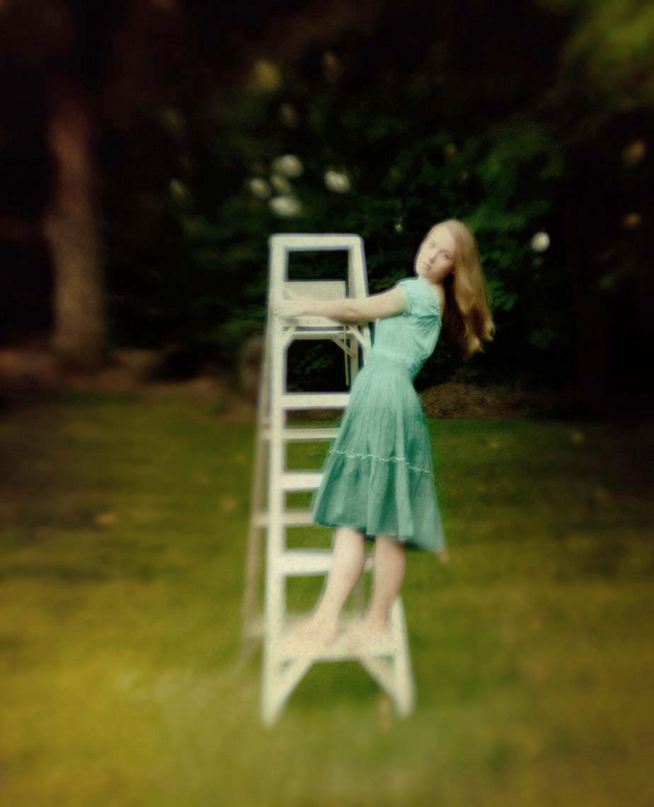 Pretty teen with long blonde hair in summer dress in blue tones standing on a step ladder on lawn with dense trees in backgroundjessica-ladder