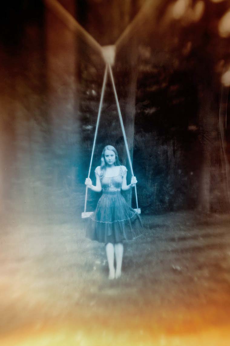Pretty teen with long blonde hair in summer dress in blue tones standing against tree swing on lawn with dense trees in background