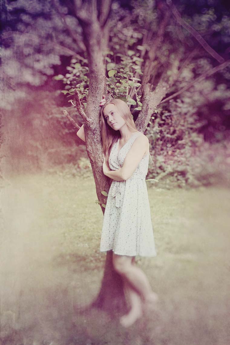Portrait of a pretty, teenage girl in a pale sleeveless summer dress, leaning against a tree, while gazing dreamily off-camera against a soft-focus landscape background.-tree