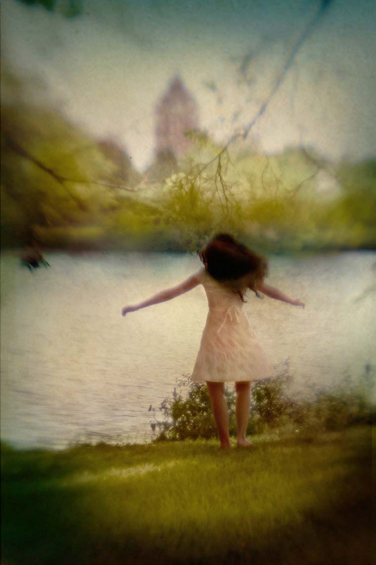 Teen girl with long brown hair in white dress, arms outstretched, twirling herself by lake in park
