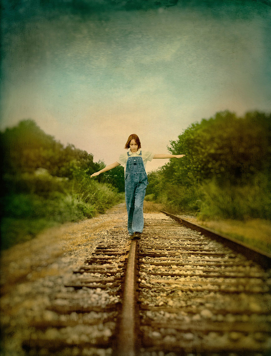 Carefully balancing on a railroad track with hands out to her sides, an adolescent girl in blue jean overalls slowly makes her way towards the viewer, while the tracks fade behind her into a melancholy landscape.