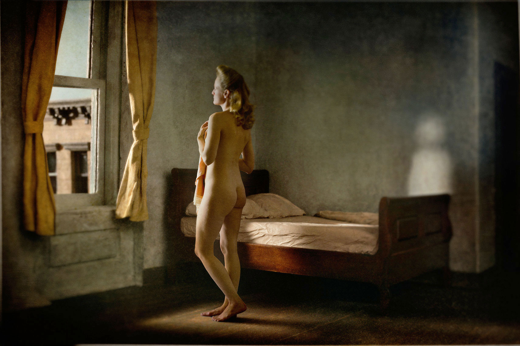 A photomontage homage to Edward Hopper’s painting, “Morning in a City”, set in a 1940s’ New York apartment bedroom, a nude melancholic blonde woman, covering the front of her torso with a garment, and standing next to an unmade bed, gazes in contemplation out the window onto the urban landscape.