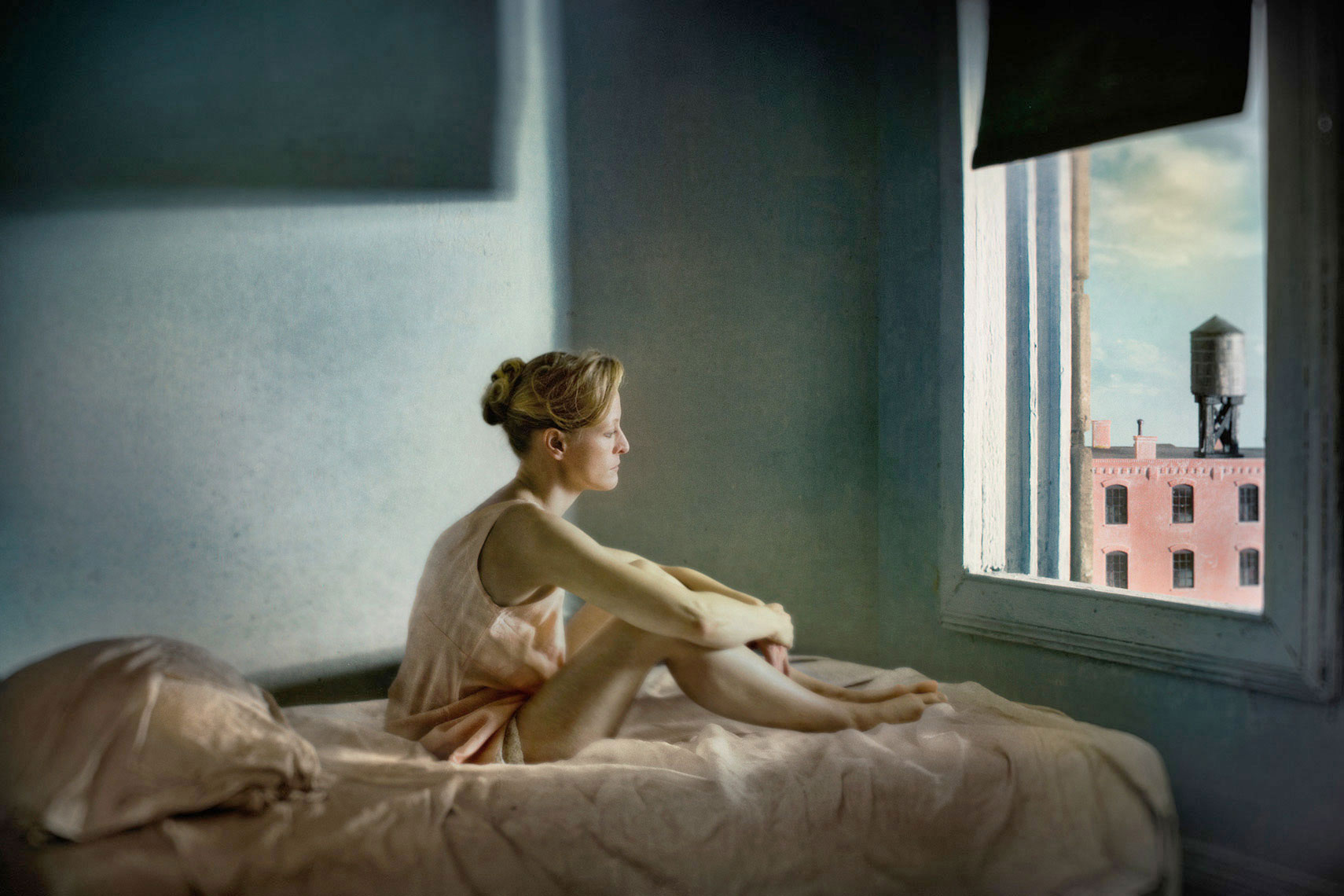 A photomontage homage to Edward Hopper’s painting, “Morning Sun”, set in a 1940s’ New York apartment bedroom, a melancholic blonde woman wearing a pale pink slip sits on the rumpled sheets of an unmade bed, gazing in contemplation out the window onto the urban landscape.