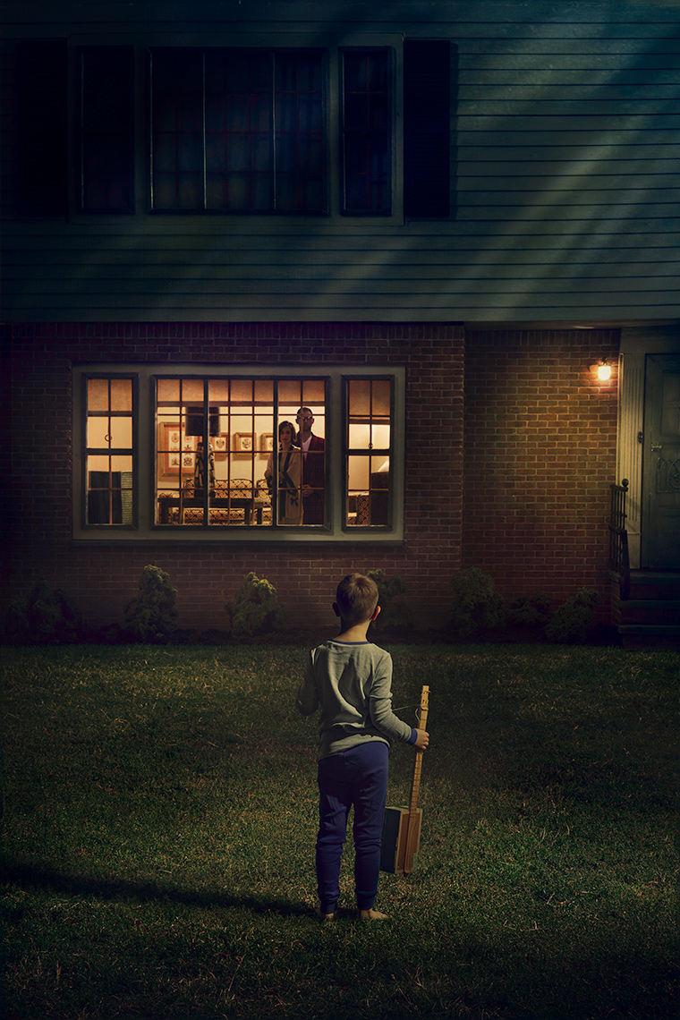 A young boy in pajamas, holding a toy guitar, stands on the lawn of a suburban house at night, seen from behind in the foreground, he stares at the front picture window, where the small figures of his parents stare back at him from inside the house.