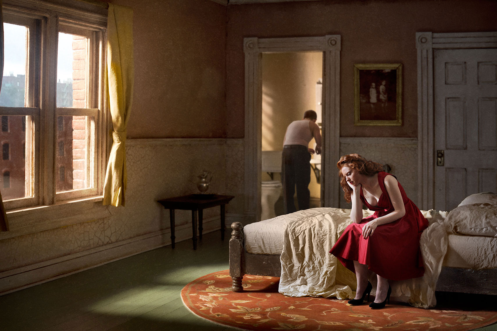 Young red-haired woman in red dress, sits on an unmade bed in a sunlit 1940s’ New York apartment while her male companion is in undershirt at the bathroom sink in the background.