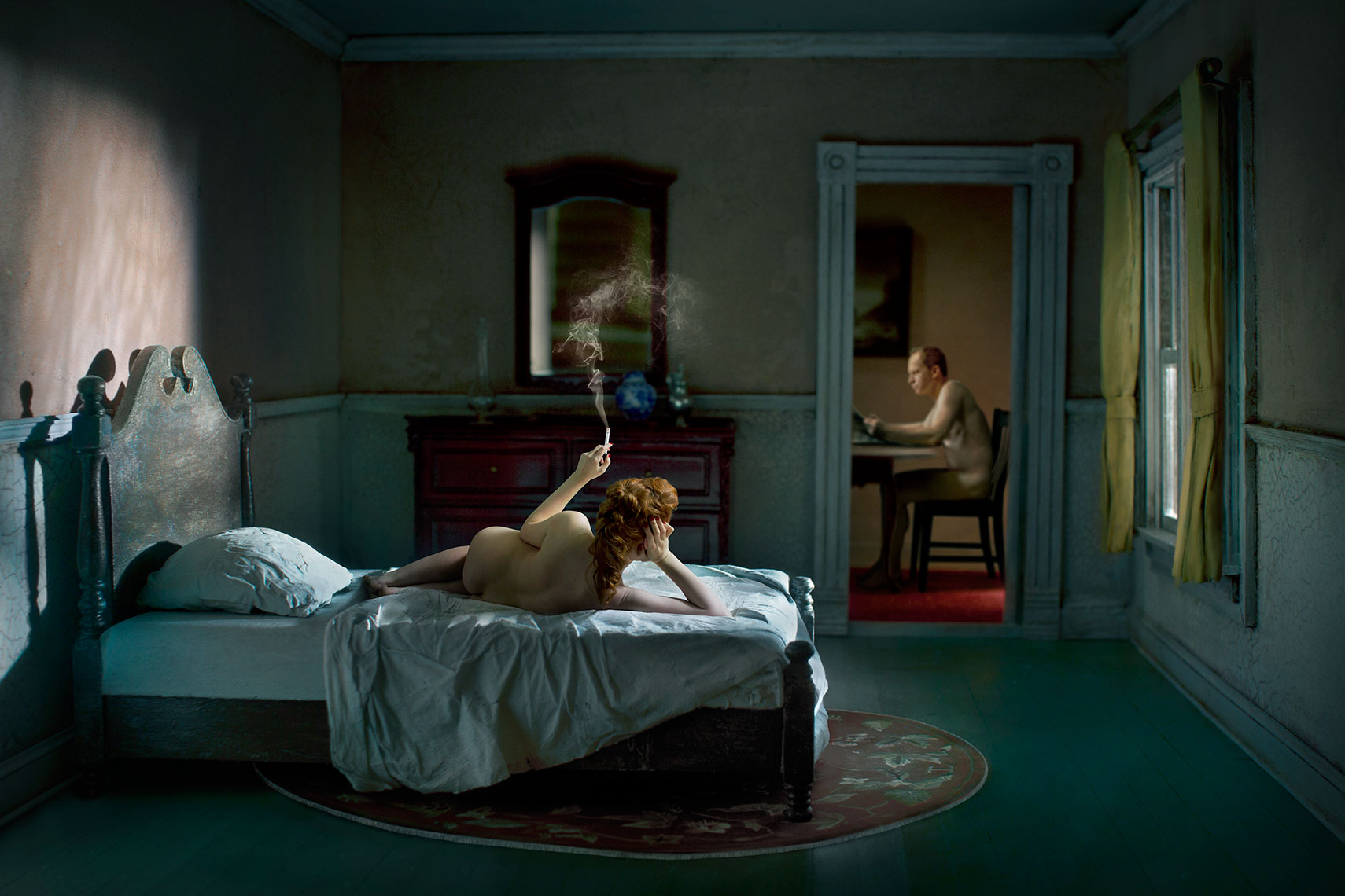 In a photomontage homage to Edward Hopper, in the foreground a young red-haired woman seen from the back lies nude on an unmade bed while smoking a cigarette in a 1940s’ New York apartment while in the background while in the background in the next room her nude male companion sits at at table reading.