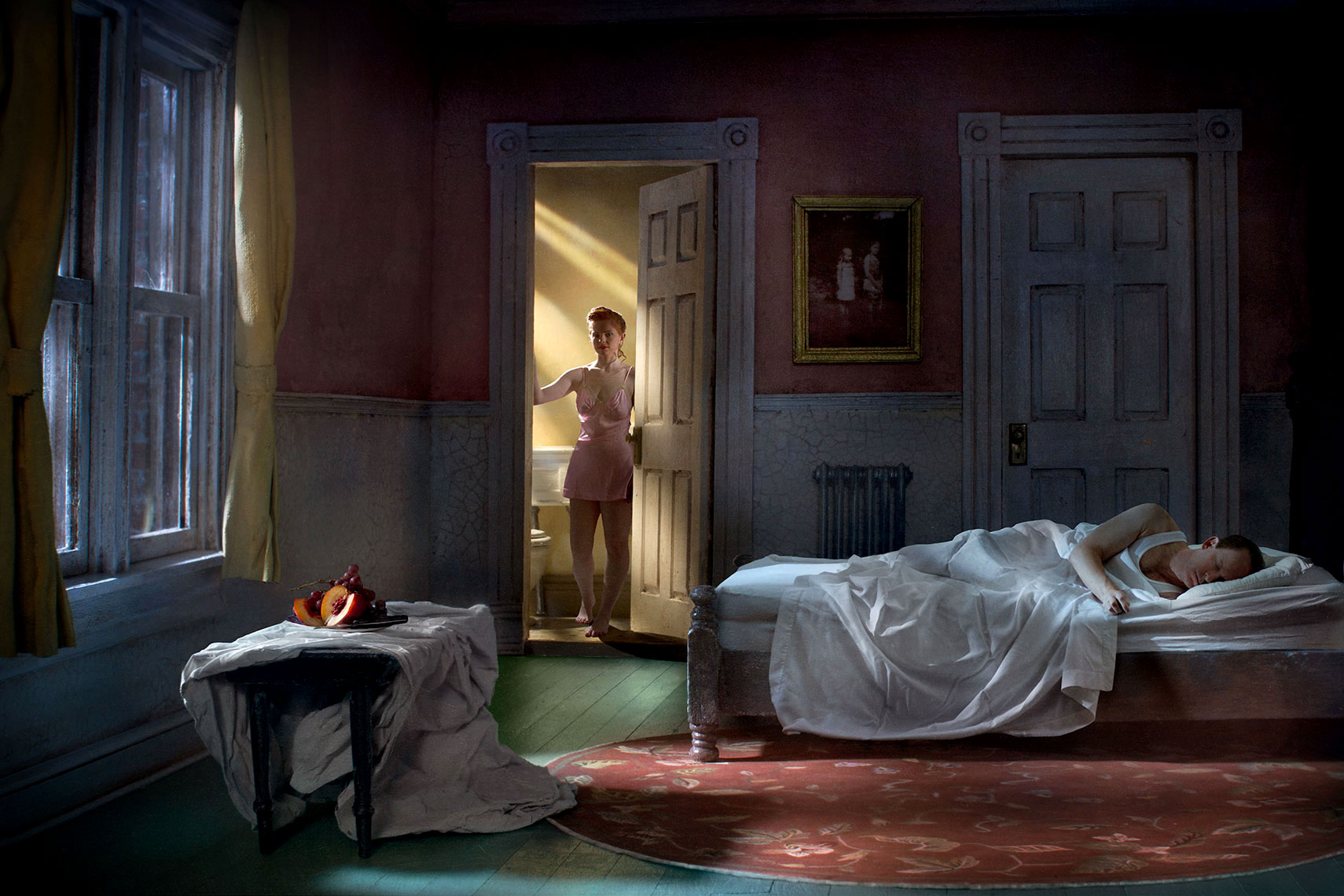 1940s’ New York apartment bedroom at night, a young woman wearing a slip emerges backlit from the bathroom in the background while her partner sleeps in the bed in the foreground, and a mysterious still life sits on a piano bench by the window, glowing in the moonlight.