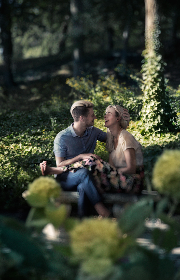 An attractive, romantic young couple sitting on a bench in a tree-filled park laughing.