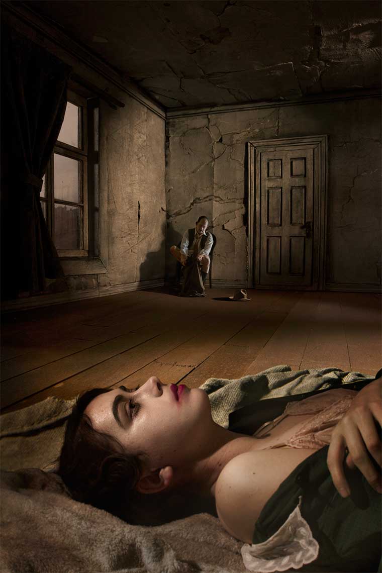 Moody painterly photomontage of a 1930s’ dingy, unfurnished decaying European apartment interior where in the foreground a beautiful dark-haired young woman, partially disrobed and makeup smeared, lies on the floor staring at the ceiling, while in the background a man sits on a chair putting his pants on.