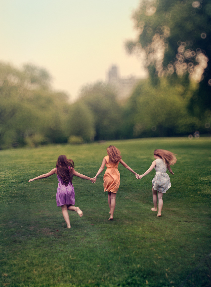 Three young women, holding hands and with hair flowing, run away from the camera toward the distant trees on the horizon of a green urban park.