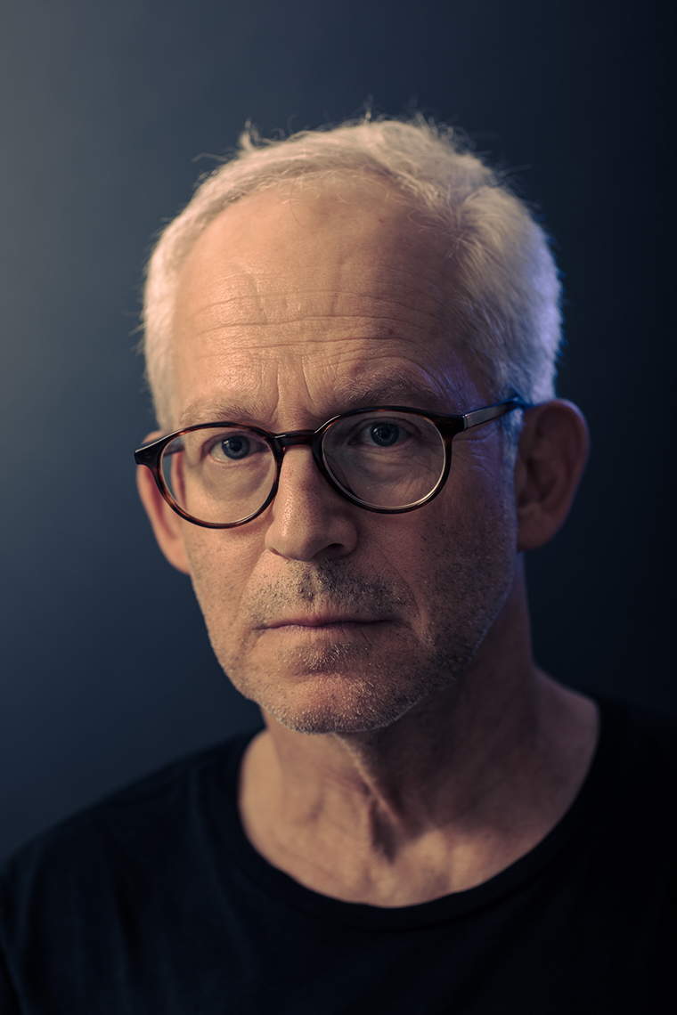 Moody portrait of older white male with short white hair, horn-rimmed glasses and a black t-shirt against a dark background.