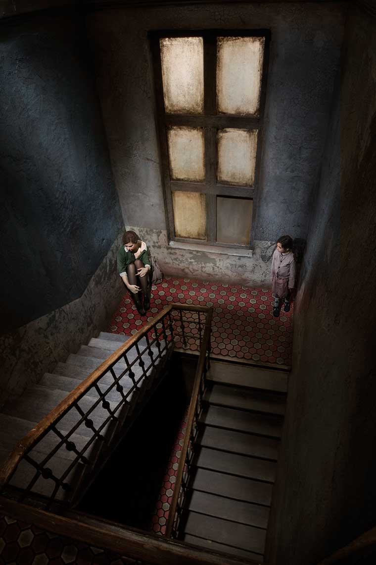 Bird’s eye view of a stairwell of a 1930s’ dingy,decaying European apartment building, where a disheveled young woman sits disconsolately on the floor in the corner of the landing, while a young girl in shabby clothes looks on.