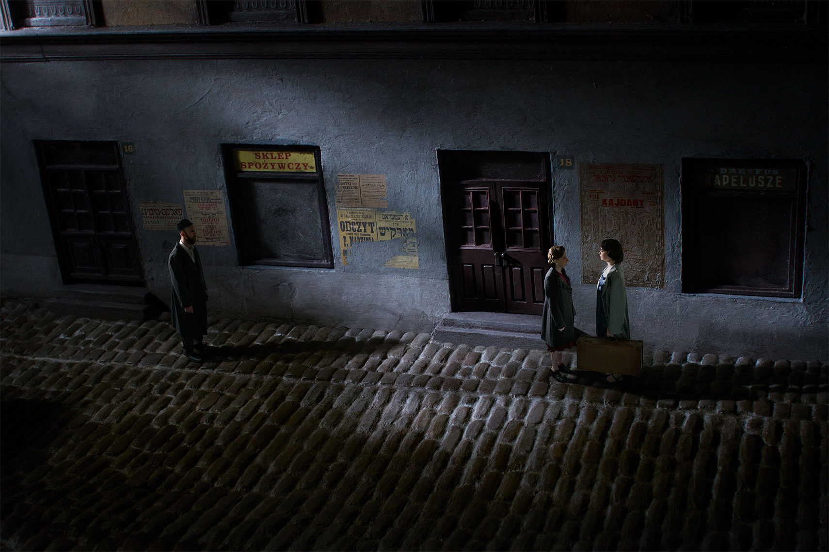 On a shadowy, dark, lonely Polish cobblestone street at night in 1930, two women, one carrying a suitcase, face each other in conversation, while ten feet away a bearded Orthodox Jewish man watches.