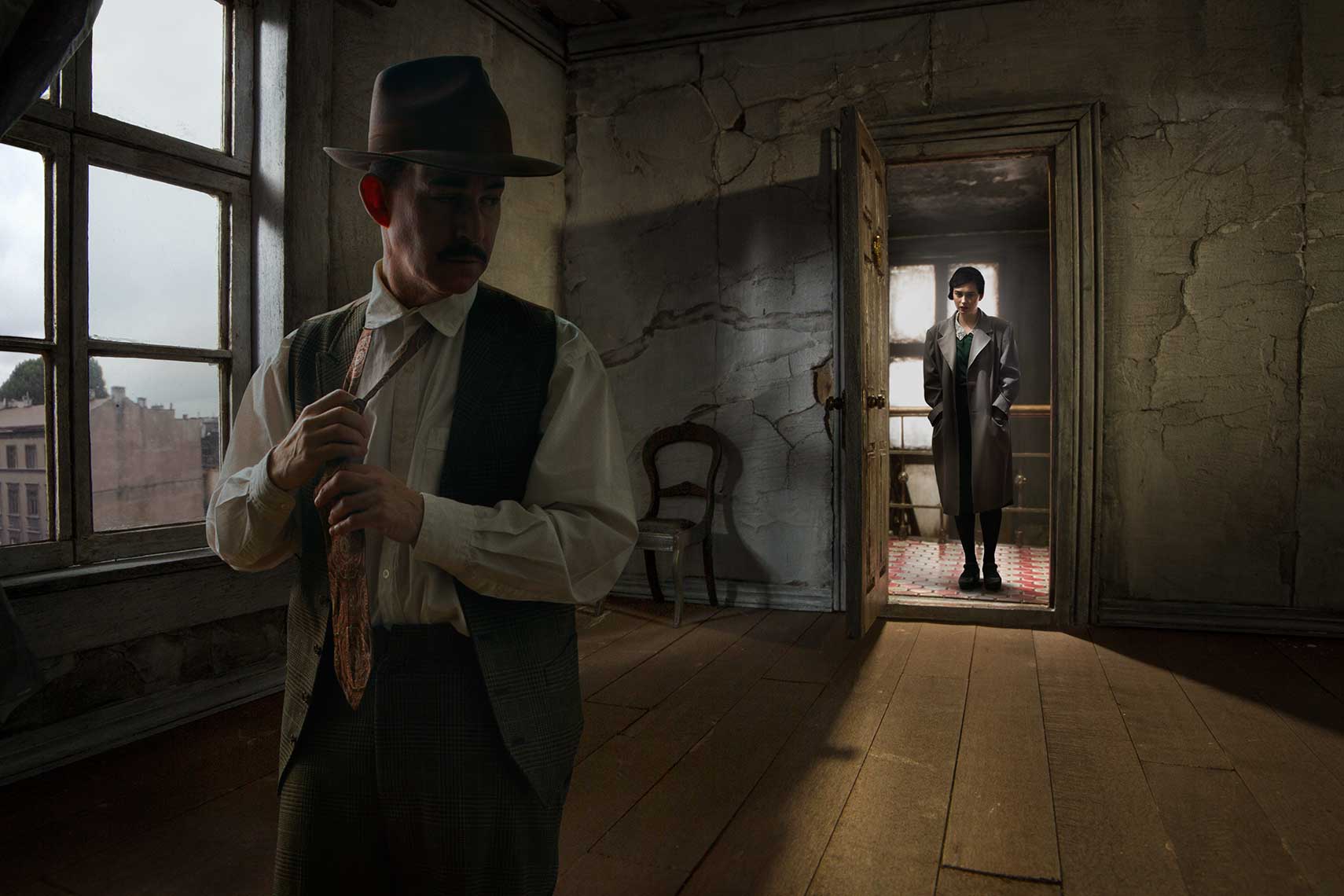 Moody painterly photomontage of a 1930s’ dingy, unfurnished decaying European apartment interior where in the foreground a shadowy male figure wearing a fedora removes his tie and turns  to look behind him at a young woman in a trench coat facing him, standing backlit in the threshold of the open door.