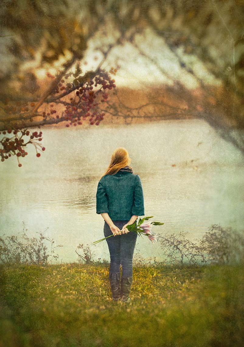 Young woman with long red hair in short jacket and jeans seen from behind standing in front of a lake in a park holding flowers behind her back.