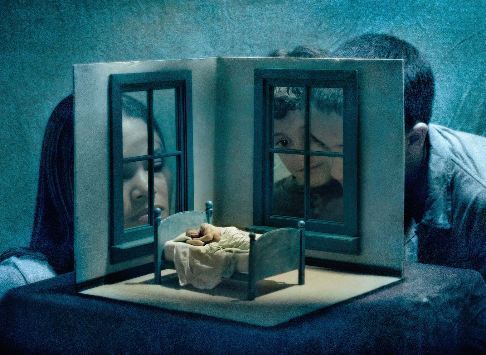 Mother and two young sons look through the windows of a dollhouse size set at the father, a miniature figure lying solemnly in a miniature bed.