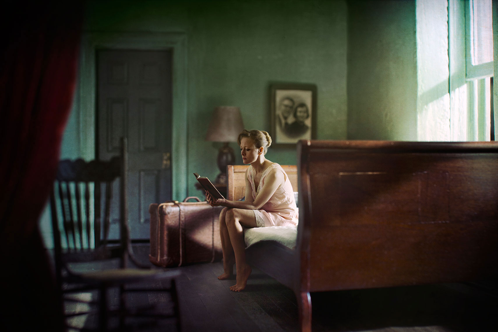 A photomontage homage to Edward Hopper, set in a 1940s’ New York apartment,  a dishwater-blonde woman wearing only a slip, sits on the edge of an unmade sleigh-bed reading a book by the bright sunlight from the open window, a suitcase standing upright on the floor next to her.