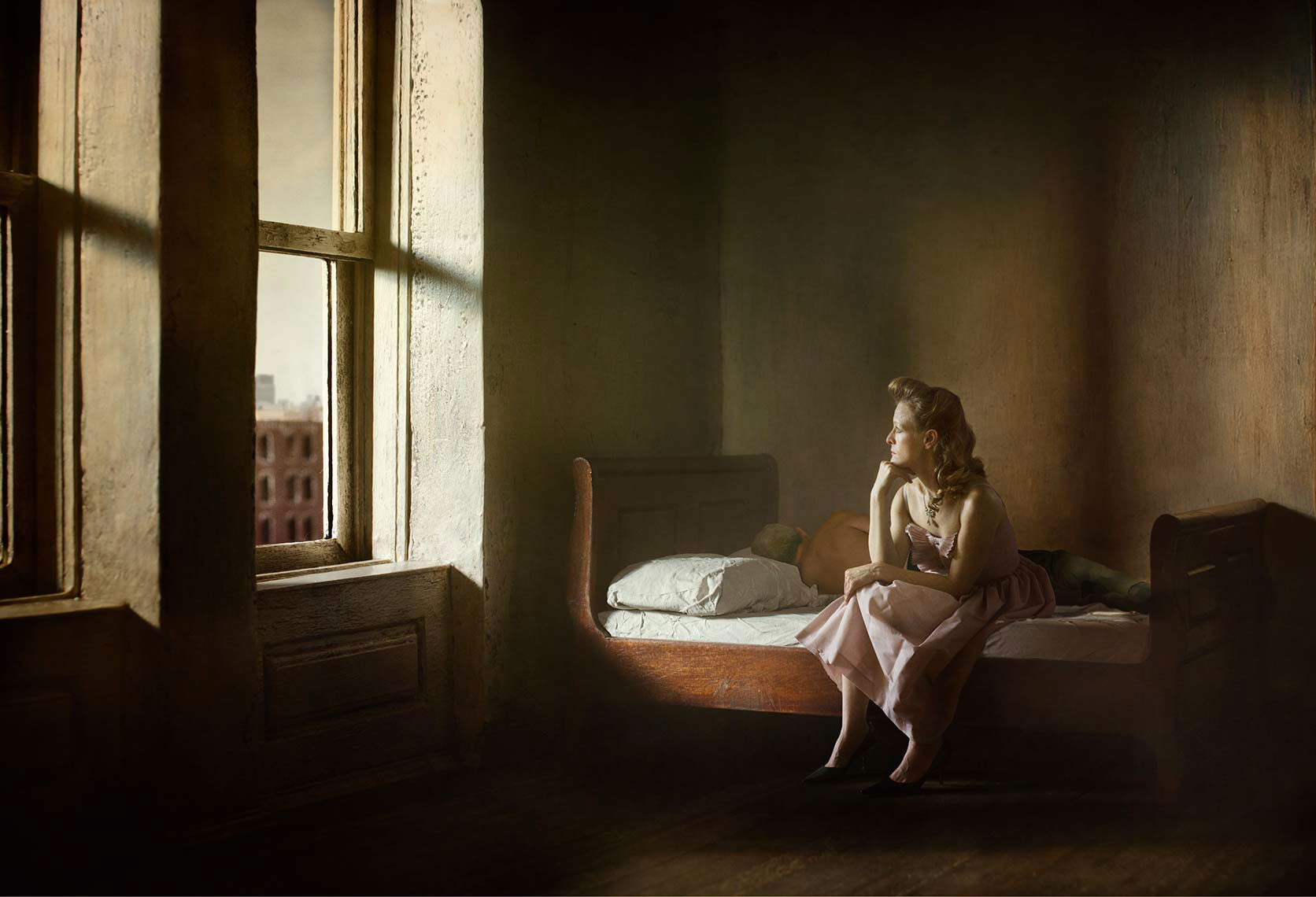 A photomontage homage to Edward Hopper’s painting, “Summer in the City”, set in a 1940s’ New York apartment bedroom, a melancholic blonde woman wearing a pale pink sleeveless dress sits on the rumpled sheets of an unmade bed, gazing in contemplation out the window onto the urban landscape, her male companion asleep in the shadows next to her.